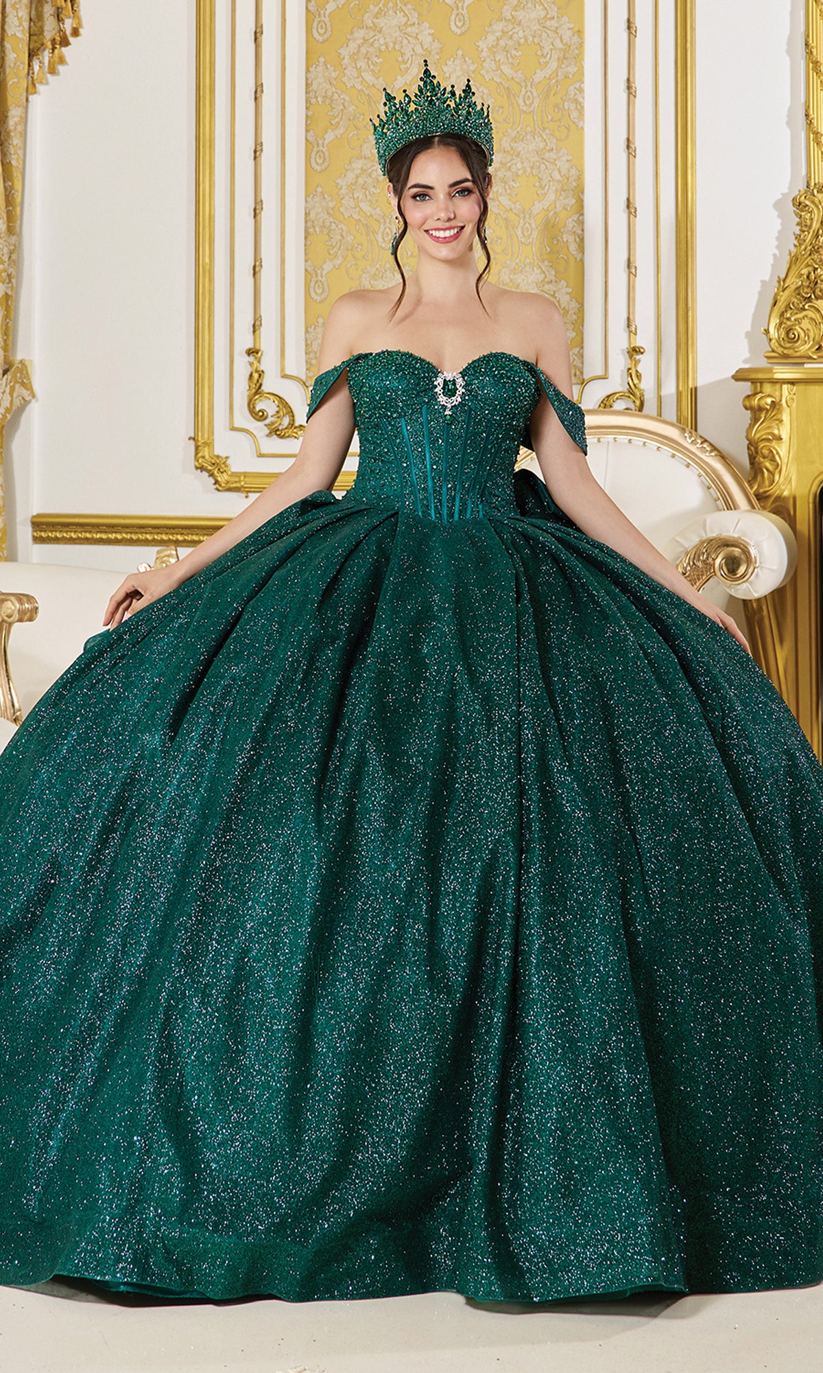 A woman in the Quinceanera Dress 1937 by Dancing Queen—a sparkling emerald green glitter ball gown with off-the-shoulder straps—wears a matching crown and stands in an ornate gold and white room.